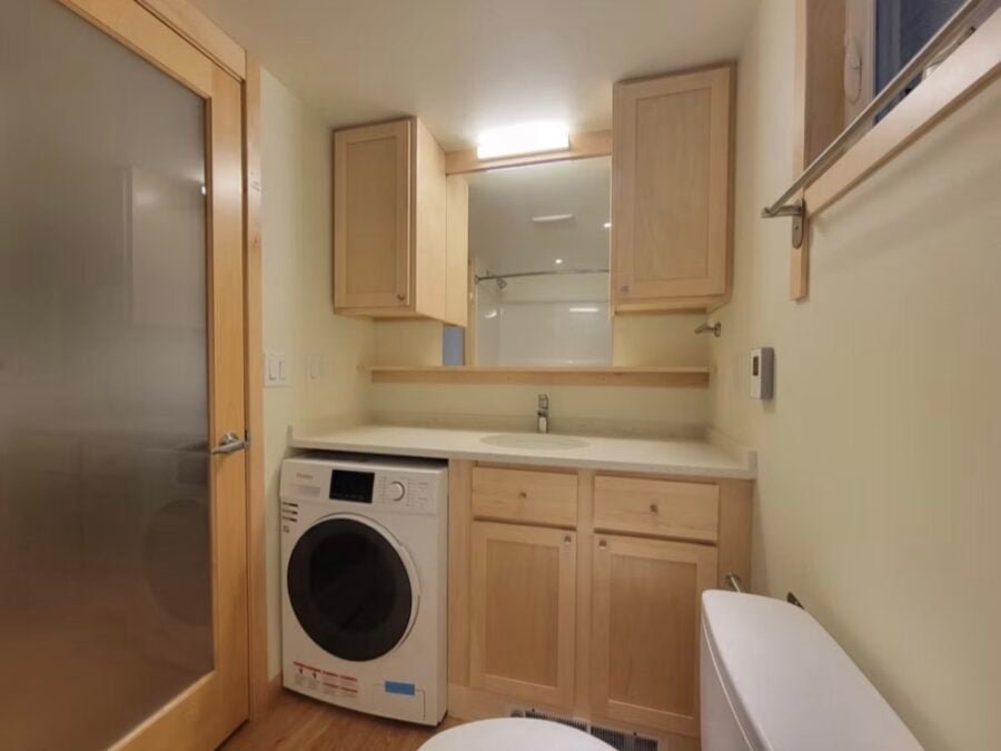 Spacious bathroom in The Grove Classic King XL Tiny House featuring a combo washer-dryer, built-in storage, flush toilet, and frosted glass door